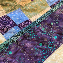 Load image into Gallery viewer, Close-up of a handmade patchwork quilt with various fabric panels, including purple and teal batik, blue starry fabrics, and beige floral prints.
