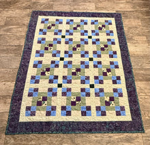 Load image into Gallery viewer, A handmade quilted blanket with a geometric block pattern in purple, blue, green, and cream, finished with a dark purple border, laid flat on a brown wood floor.
