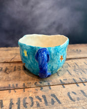 Load image into Gallery viewer, A handmade ceramic cup with a blue handle and yellow star details, sitting on a rustic wooden surface with a smoky gray background.
