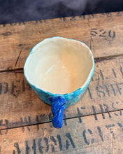 Load image into Gallery viewer, A handmade ceramic mug with a textured turquoise blue exterior and a glossy cream-colored interior, featuring a sculptural blue handle shaped like a small animal head, resting on a rustic wooden surface.
