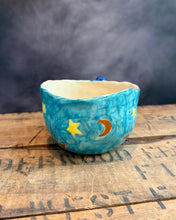 Load image into Gallery viewer, A small handmade ceramic cup with a blue sky and moon design, sitting on a rustic wooden surface with a smoky dark background.
