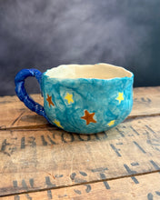 Load image into Gallery viewer, A handmade ceramic cup with a marbled blue exterior, yellow star patterns, and a dark blue handle, resting on a rustic wooden surface with a smoky gray background.
