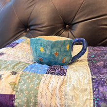 Load image into Gallery viewer, A handmade blue ceramic mug decorated with yellow stars and orange crescent moons, resting on a colorful patchwork quilt in front of a dark brown tufted leather headboard.
