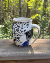Load image into Gallery viewer, White mug with black and blue patterns on a wooden surface with a forest background

