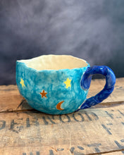 Load image into Gallery viewer, A handmade ceramic cup with a blue sky and starry night theme, featuring a textured turquoise body, a dark blue handle, and yellow stars and a crescent moon, resting on a rustic wooden surface.
